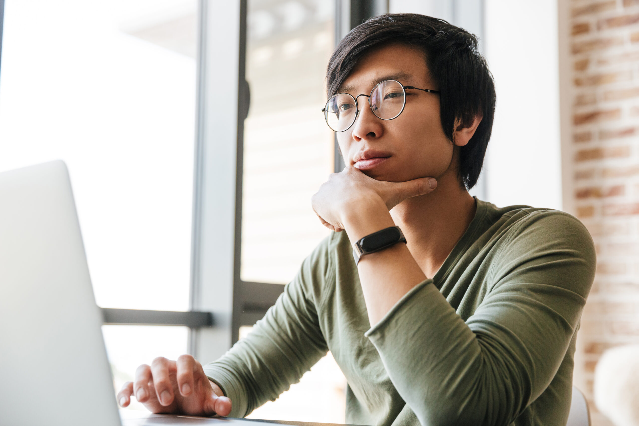 Image of handsome young asian man using laptop in apartment Image of handsome young asian man wearing eyeglasses using laptop while sitting at table in apartment