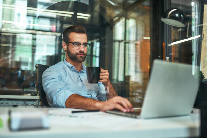 Morning coffee. Portrait of young and successful bearded man in eyeglasses holding cup of coffee and working with laptop while sitting at his working place Morning coffee. Portrait of young and successful bearded man in eyeglasses holding cup of coffee and working with laptop while sitting at his working place. Business concept. Work concept
