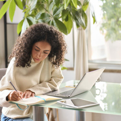 Young woman student using laptop watching online webinar writing at home. Young woman, girl student using laptop elearning or remote working at home office using laptop pc computer for webinar, learning online training digital education sitting at table, writing notes.