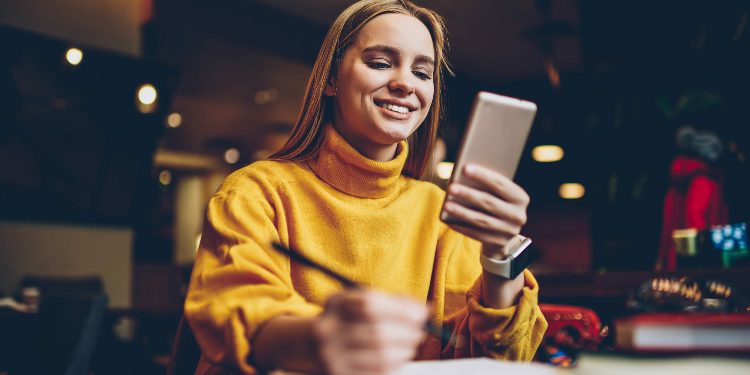 Pretty prosperous blonde woman dressed in cool yellow sweater using smartphone application for planning working schedule.Cute successful female blogger chatting with followers in social network