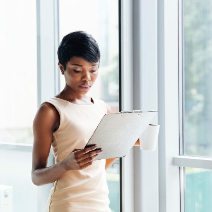 Beautiful african young businesswoman holding clipboard and reading in office Beautiful african american young businesswoman with cup of coffee holding clipboard and reading in office